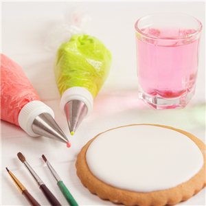 Two tubes of frosting, one pink and one green, a cup of pink tinted water, three paintbrushes, and a sugar cookie with white frosting. 