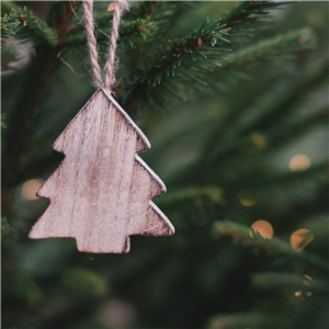 A close up picture of a wooden tree shaped Christmas ornament hanging on a lit branch of a tree