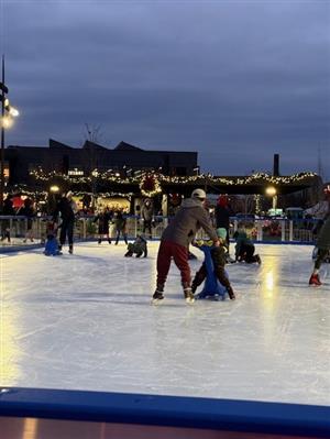 A dad and son ice skating on the ice rink with holiday lights and a lit wreath in teh background.