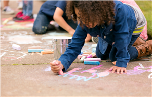 A young child with curly hair creating chalk designs on a sidewalk.