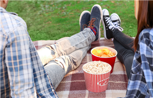 Two people sitting on a picnic blanket in the grass with a bucket of popcorn between them.